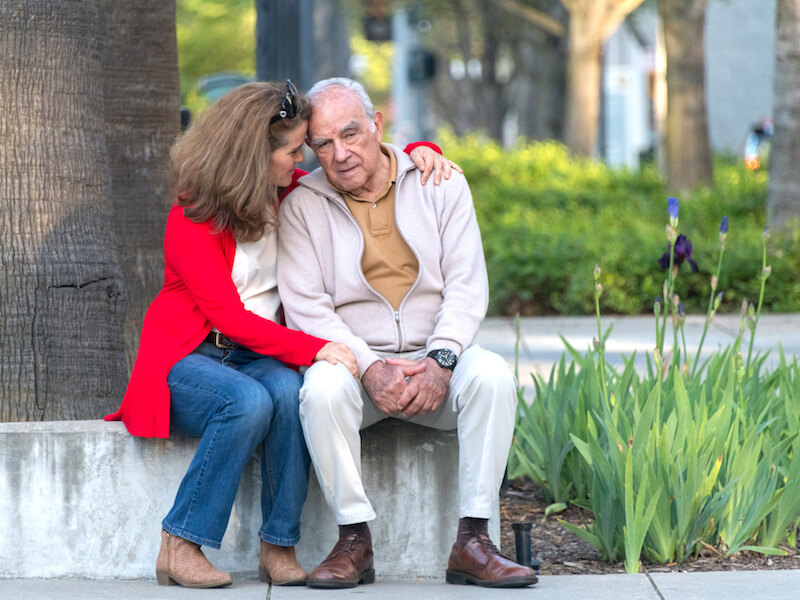 A woman in a red sweater embraces an elderly man in a light jacket as they sit on a stone bench. They appear engaged in a tender conversation. Greenery and flowers are visible in the background.