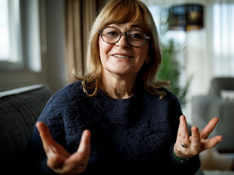 A smiling woman with glasses and light brown hair sits on a couch, gesturing with her hands. Shes wearing a dark sweater, and the background features a window with light coming through and a blurred indoor plant.