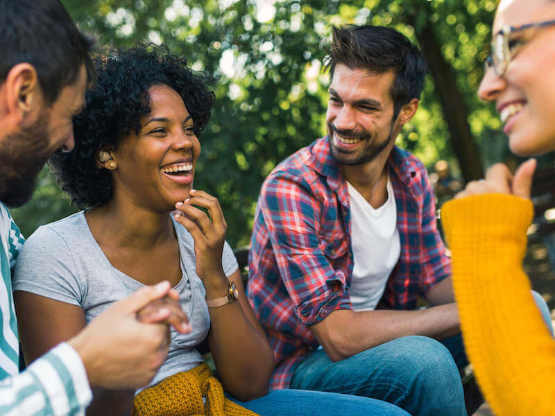 Four people are sitting outside, laughing and talking. One wears a gray shirt, another a plaid shirt, and another a mustard sweater. They seem to be enjoying a lively conversation in a green, leafy setting.