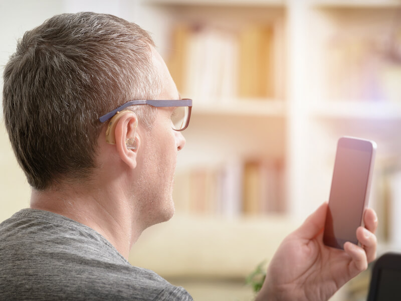A man with glasses and a hearing aid looks at a smartphone. He is wearing a gray shirt, and the background features a blurred bookshelf. The setting appears to be indoors, possibly a home or office.