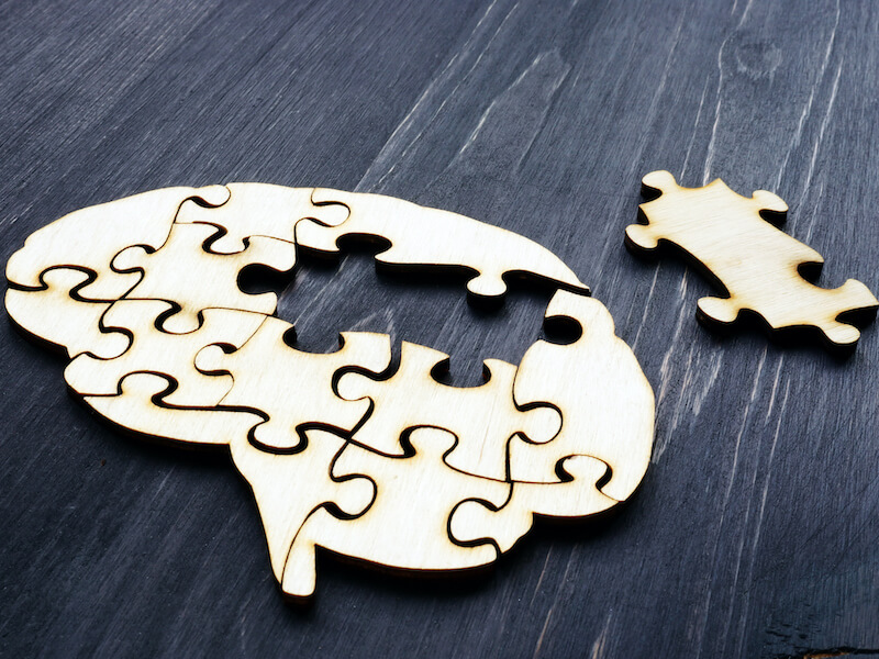 A wooden puzzle shaped like a human brain is nearly complete on a dark wooden surface. One puzzle piece is placed outside, ready to fit into the remaining hollow space within the brain shape.