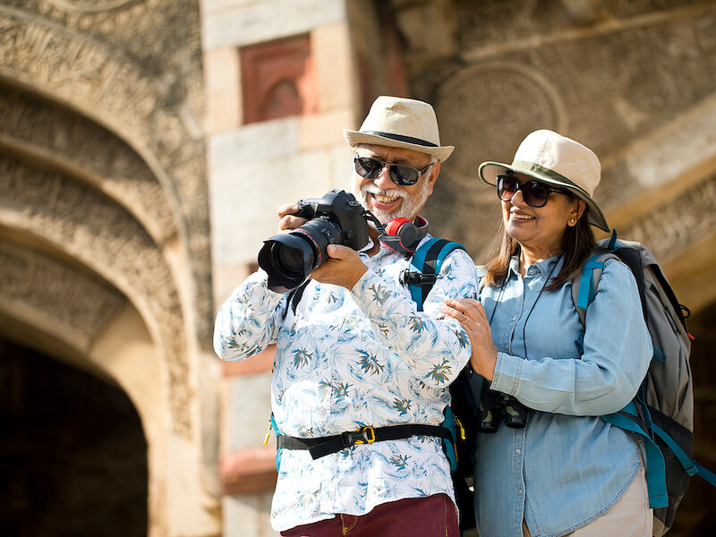 An older couple with hats and sunglasses happily looking at a camera screen. The man wears a floral shirt and red pants, while the woman wears a blue shirt. They are in front of an ancient architectural structure. Both have backpacks.