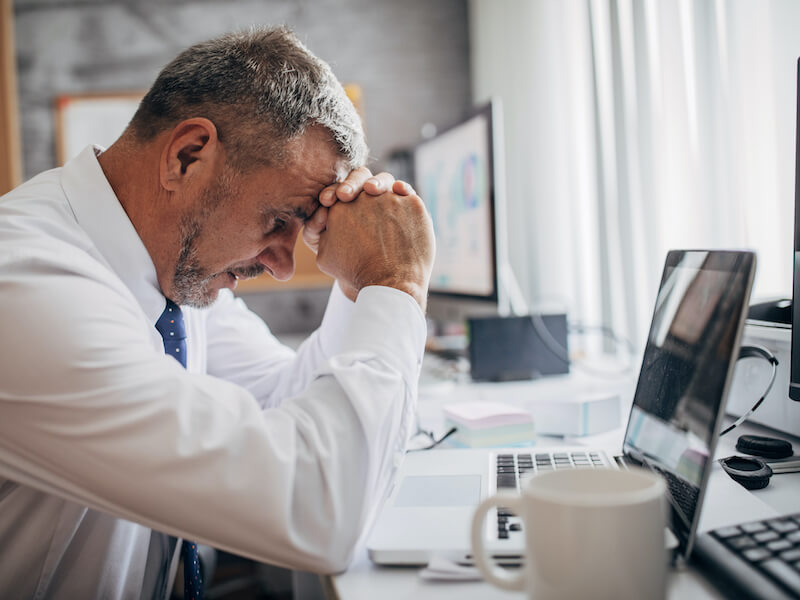 A man in a white shirt and tie sits at a desk, resting his forehead on his clasped hands in front of a laptop. The background includes computer monitors and office supplies, and a coffee mug is in the foreground.