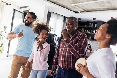 A group of four people smiling and having fun indoors. They are standing in a living room, holding microphones, suggesting a karaoke session. The atmosphere is joyful and lively, with a modern kitchen visible in the background.
