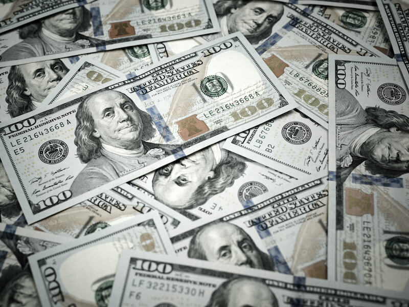 A pile of U.S. one hundred dollar bills featuring Benjamin Franklin. The bills are spread out and overlapping, showing the front side with green and blue tones.