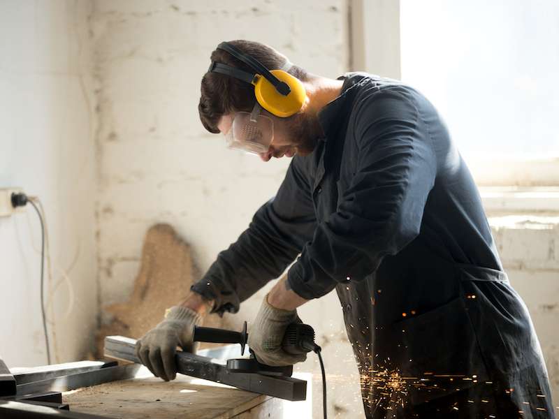 A man in protective goggles and earmuffs uses an angle grinder on a metal piece. Sparks fly as he works in a bright workshop, wearing gloves and a dark outfit. A window on the right lets in natural light.