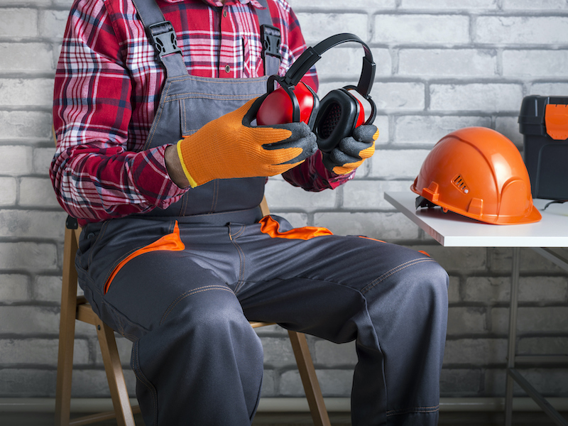 A person in work overalls, orange gloves, and a red plaid shirt sits on a chair holding noise-cancelling headphones. A hard hat and toolbox are on the table beside them, with a brick wall in the background.