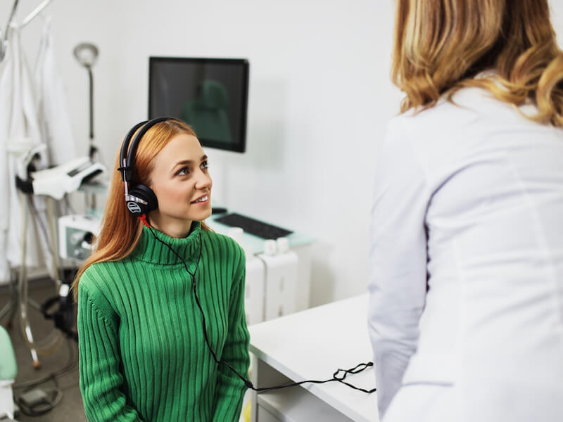 A woman with long red hair wearing a green sweater and headphones sits in a medical office, looking at a healthcare professional in a white coat. Behind them is medical equipment and a computer monitor.