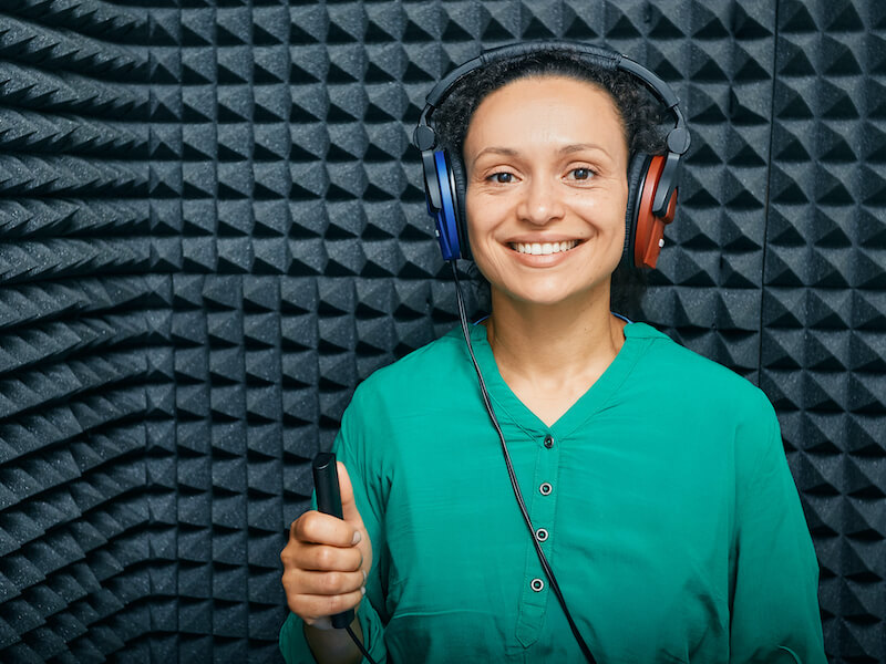A person with headphones smiling in a soundproof, foam-lined room, holding a microphone. They are wearing a green shirt and appear to be in a recording studio.