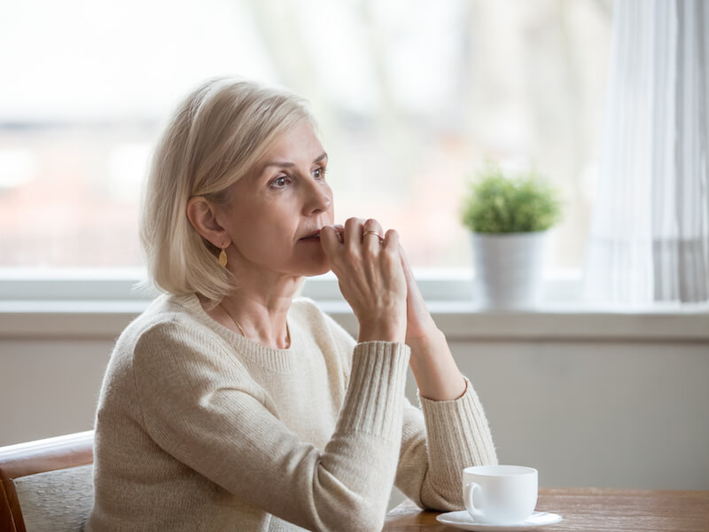 A woman with short blonde hair sits at a table, wearing a beige sweater. She looks thoughtful, resting her chin on her hands. A small white cup and a potted plant are on the table in front of her, and a window is in the background.