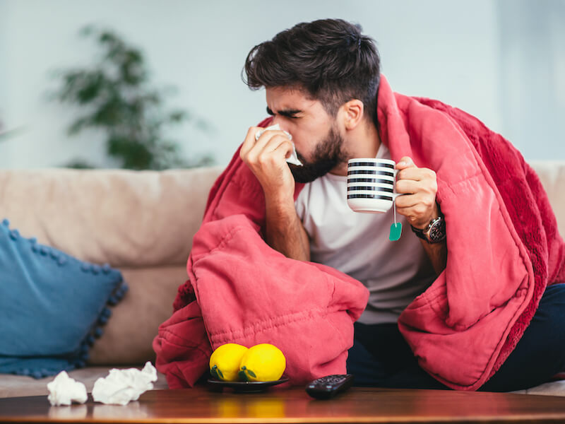 A man sits on a couch wrapped in a red blanket, holding a striped mug and blowing his nose with a tissue. There are lemons and crumpled tissues on the table in front of him. A blue pillow is on the couch beside him.