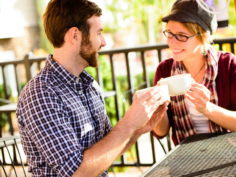A man and woman sit at an outdoor cafe table, smiling at each other while holding white coffee cups. The man wears a plaid shirt, and the woman wears glasses and a hat. A metal fence and greenery are in the background.