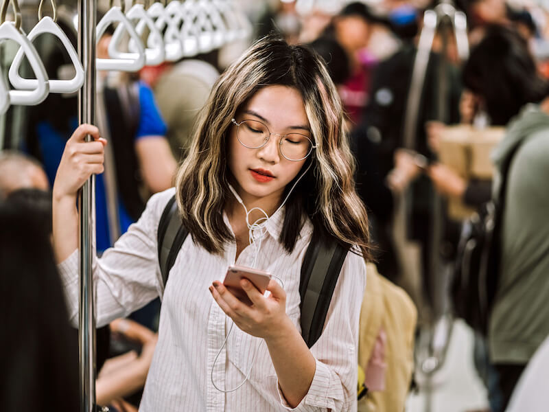 A woman with glasses and headphones is looking at her smartphone on a crowded subway. She has a backpack and holds onto an overhead handle. The subway is filled with standing passengers.