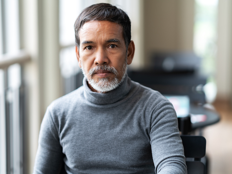 A person with a beard and mustache wearing a gray turtleneck sweater sits indoors, looking directly at the camera. The background is softly blurred, showing large windows and some furniture.