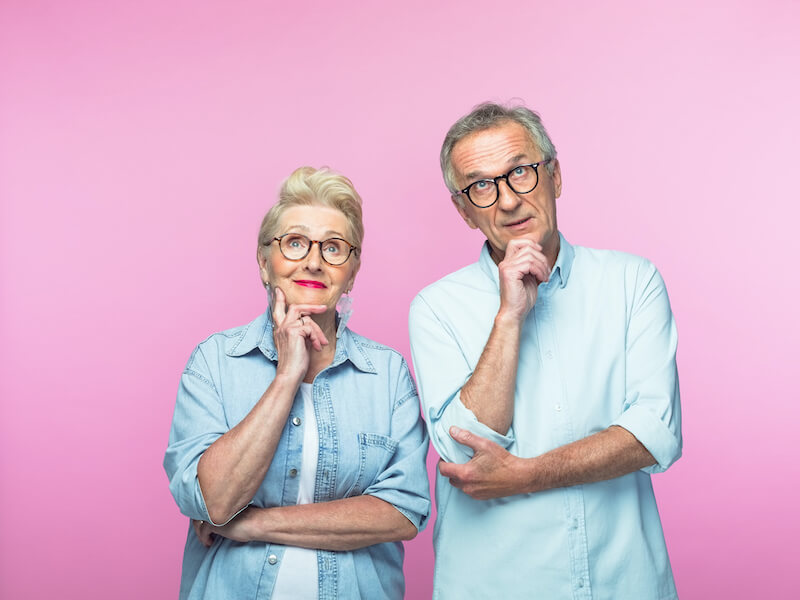 An elderly man and woman stand against a pink background, both wearing glasses and casual blue shirts. They have thoughtful expressions, with their hands resting on their chins.