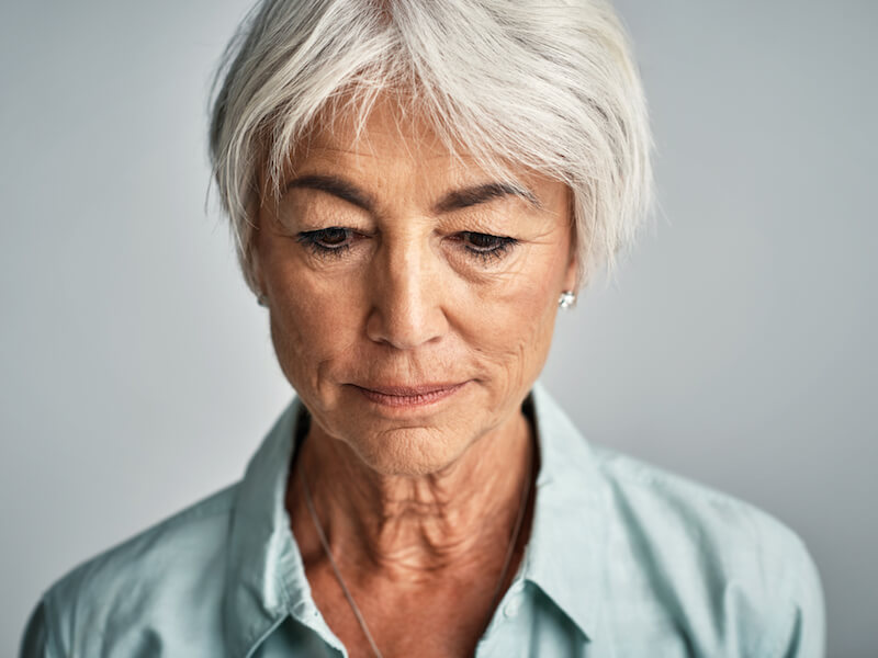 An older woman with short gray hair looks down, wearing a light blue collared shirt. Her expression is contemplative, and she stands against a plain background.