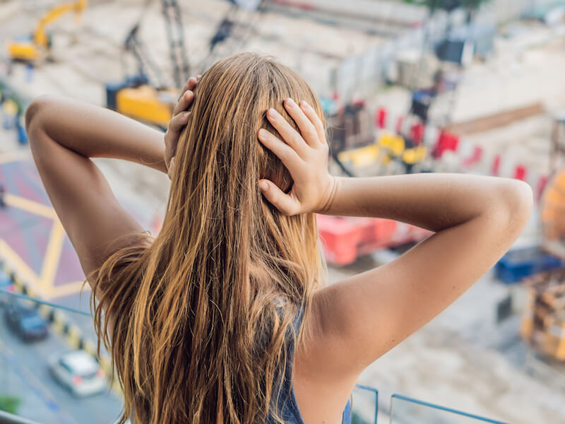 A woman with long hair stands on a balcony, covering her ears with her hands. She is overlooking a construction site with cranes and machinery in the background.
