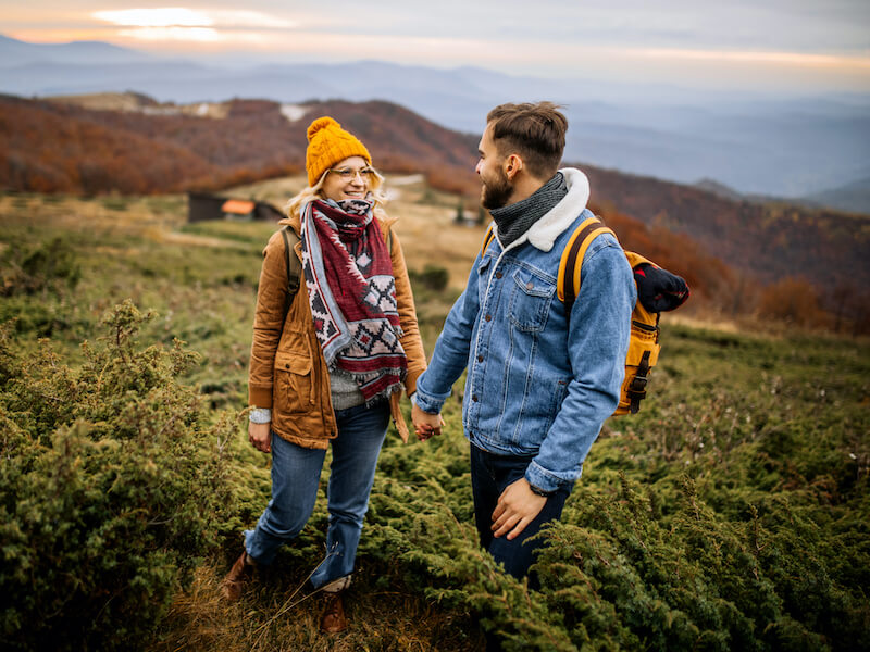 A couple stands hand-in-hand on a grassy hill, dressed warmly in jackets, scarves, and a yellow beanie. The man carries a backpack. Rolling hills and a cloudy sky are visible in the background, suggesting a chilly day.