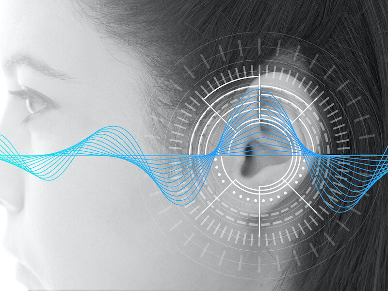 Profile of a persons face with their ear highlighted by digital graphics, including concentric circles and a soundwave pattern, suggesting technology related to hearing or sound analysis. The overall tone is monochrome with blue accents.