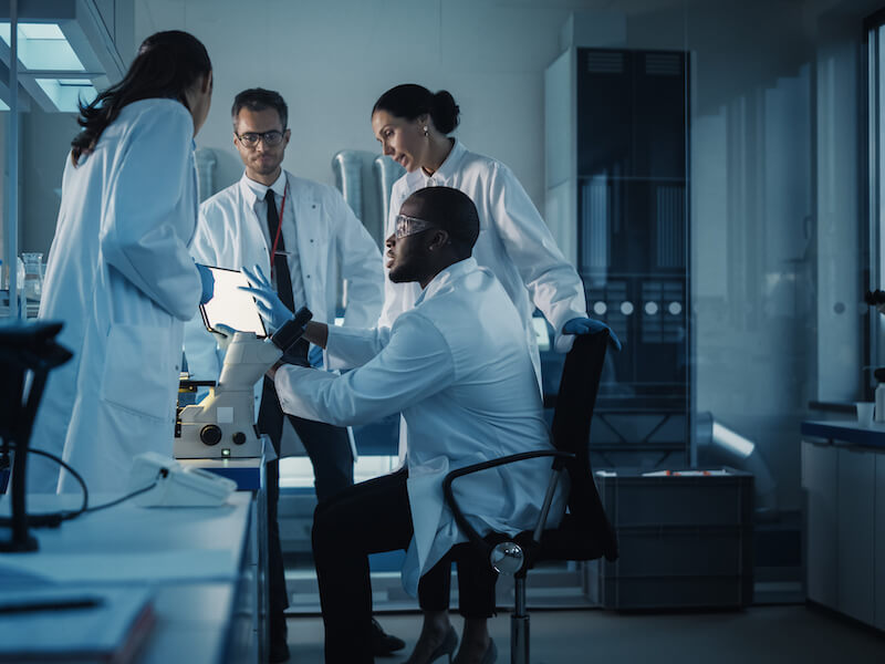 Four scientists in lab coats are engaged in discussion in a modern laboratory. One is seated, looking through a microscope, while the others stand around, examining a tablet. The lab is equipped with shelves and scientific equipment.