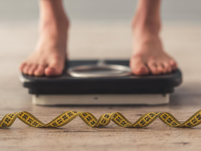 Close-up of a persons bare feet standing on a black bathroom scale. In the foreground, a yellow measuring tape is coiled, displaying numbers. The setting appears to be a wooden floor.