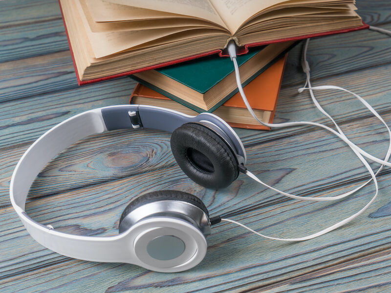 A set of silver and black headphones rests on a wooden surface, with its cable intertwined around three stacked books, including an open one on top. The books have colorful covers against the textured wooden background.
