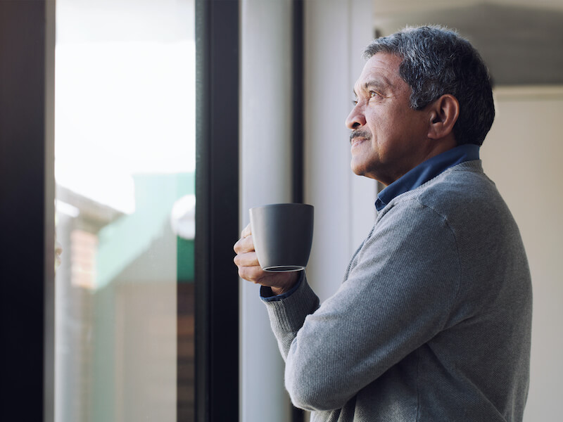 A man with gray hair, wearing a gray sweater, holds a mug while gazing thoughtfully out a window. He stands indoors, with natural light streaming in, creating a peaceful ambiance.