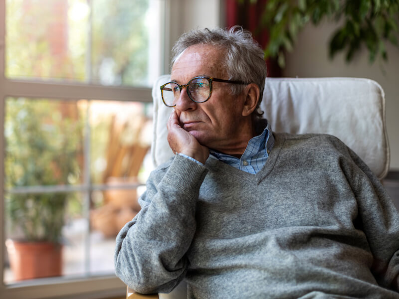 An older man wearing glasses and a gray sweater sits in a chair by a window, resting his chin on his hand, gazing thoughtfully outside. The background includes a large plant and a glimpse of outdoor greenery.