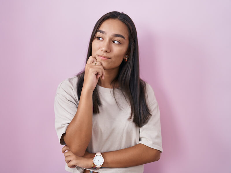 A woman with long dark hair stands against a pink background, wearing a beige shirt and a watch. She has a thoughtful expression, with one hand resting on her chin and the other crossed over her body.