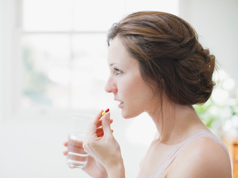 A woman with brown hair is holding a glass of water and a pill near her mouth, appearing ready to take it. She is standing indoors with a blurred window in the background.