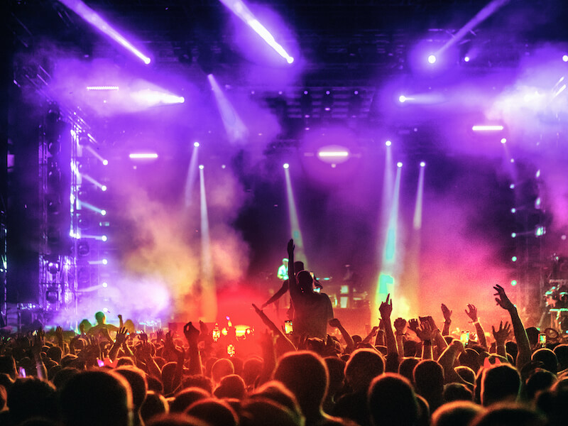 A vibrant concert scene with colorful lights illuminating a large crowd. People raise their hands and enjoy the energetic atmosphere, while smoke and bright beams create a dynamic backdrop on the stage.