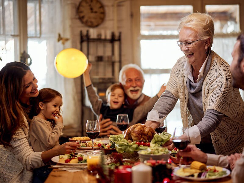 A joyful family gathers around a dinner table. An elderly woman serves a roasted turkey as others smile. Two children sit happily, and adults raise glasses of wine. The setting is warmly lit and cozy, suggesting a festive meal.