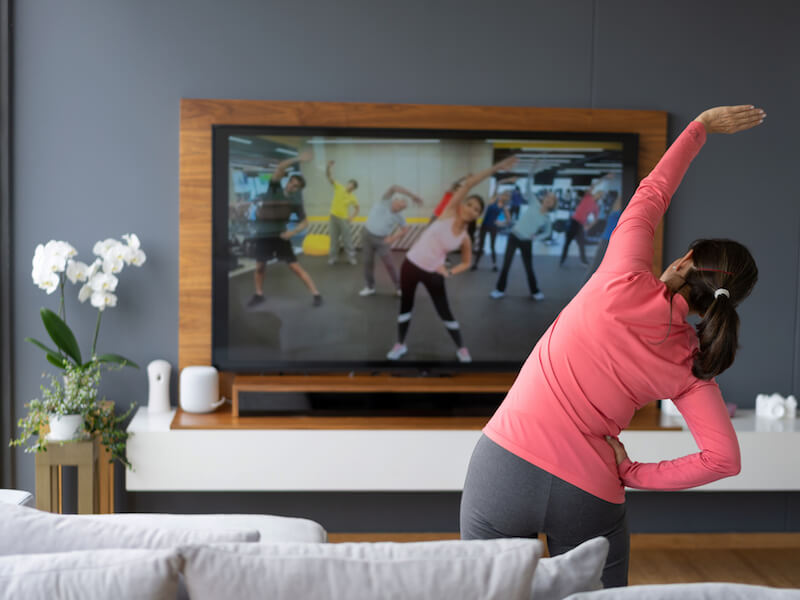 A person in a pink top and gray pants is doing a side stretch while following a group exercise video on a large TV screen in a living room. Theres a white orchid on a table next to the TV.