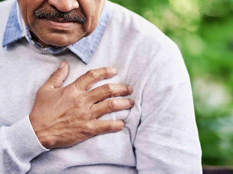 An older person with a mustache is holding their chest with their right hand, wearing a light gray sweater over a blue collared shirt. The background is blurry and green, suggesting an outdoor setting.