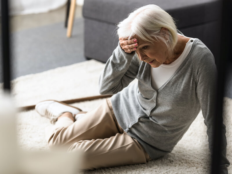 An older woman with short white hair sits on the floor with a hand on her forehead, looking distressed. She is wearing a gray cardigan, white shirt, and beige pants. A dark sofa is in the background.