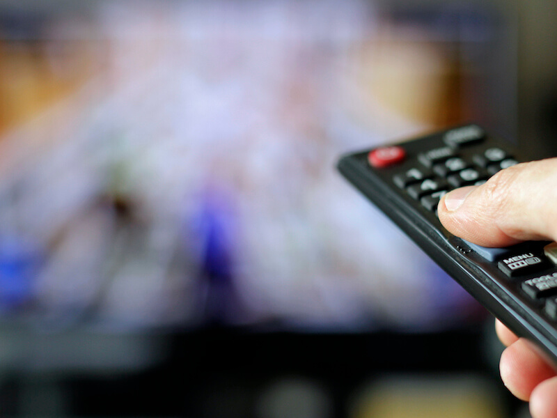 A hand holding a remote control is pointing towards a blurred television screen. The remotes buttons are visible, with a finger poised to press one. The background shows an indistinct image on the TV.