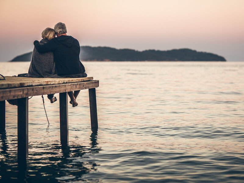 A couple sits on a wooden dock, embracing as they gaze at a calm sea during sunset. An island is visible in the distance under a soft pink and orange sky.