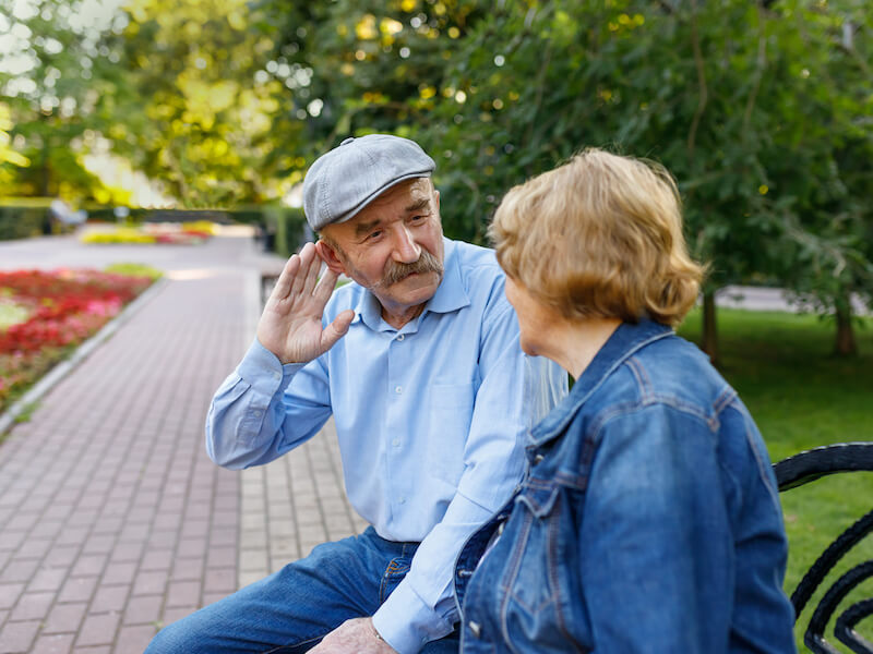 An elderly man in a cap sits on a park bench, leaning forward with a hand to his ear, listening to a woman in a denim jacket. They are surrounded by greenery and flowers on a sunny day.