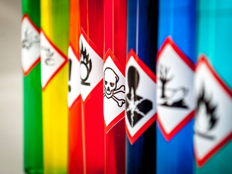 Colorful test tubes arranged in a row, each labeled with hazardous symbols, including flammable, toxic (skull and crossbones), and other warning signs, indicating chemical substances.