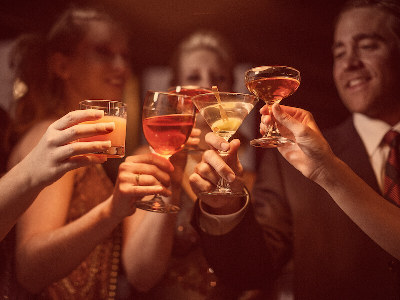 A group of people clinking various cocktail glasses in a celebratory toast. The setting appears festive with dim lighting and warm tones. Everyones attire is formal, creating a sophisticated atmosphere.