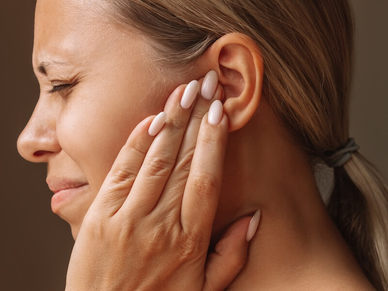 A person with long, manicured nails holds their hand to their ear, appearing to be in discomfort or pain. They have closed eyes and light brown hair tied back. The background is simple and neutral.