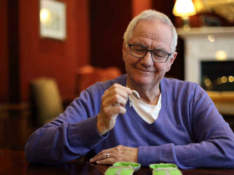 Elderly man in glasses and a purple sweater sitting at a table, examining a green object in his hand, with a smile. The background shows a cozy room with warm lighting and furniture.