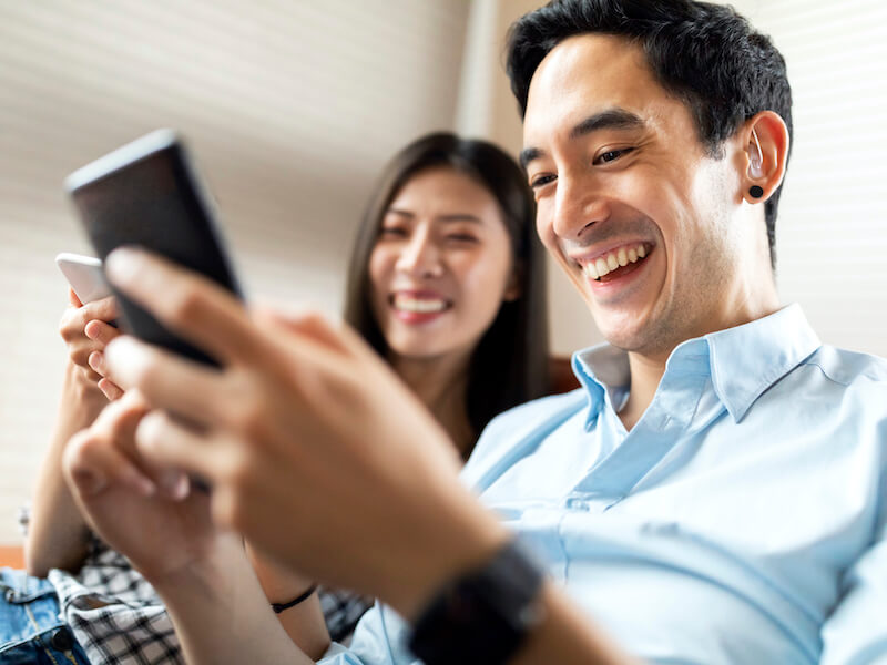 A smiling man and woman are sitting closely, each holding a smartphone. The man in the foreground appears engaged with his device, and both look relaxed and content in a bright indoor setting.