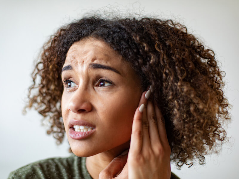 A person with curly hair looks distressed, holding their hand to their ear. They appear concerned or in discomfort, with a neutral background.