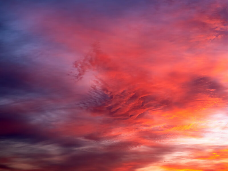 A vibrant sunset sky with swirling clouds in warm hues of red, orange, and pink, blending into cooler purple and blue tones. The cloud patterns create a dynamic and colorful scene.