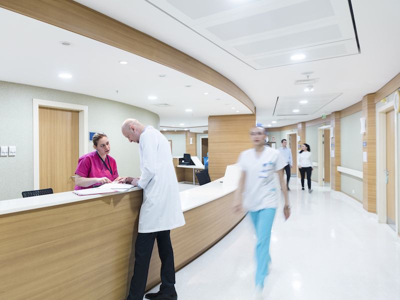 A hospital scene with a male doctor and a female nurse discussing paperwork at a curved wooden reception desk. Blurred medical staff in white coats walk through a bright, modern corridor.