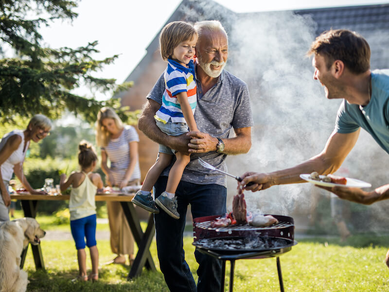 A cheerful outdoor barbecue scene. An older adult holds a young child while another man grills food on a smoky charcoal barbecue. A family, including a dog, enjoys themselves at a wooden table in the background on a sunny day.