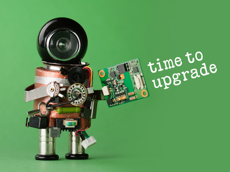 A small robot made of electronic components holds a circuit board against a green background. The robot has a large camera lens as a head. The text time to upgrade is displayed beside it.