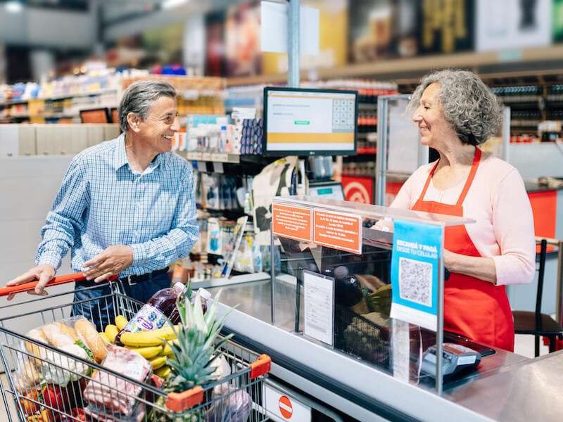 A man at a supermarket checkout smiles at a cashier wearing a red apron. His cart is filled with groceries, including a pineapple and bread. They appear to be having a friendly conversation.