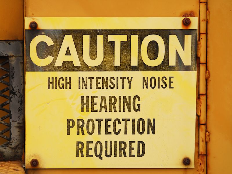 Yellow caution sign with bold black text reads: Caution. High Intensity Noise. Hearing Protection Required. The sign is posted on a yellow surface with a grated pattern visible to the left.